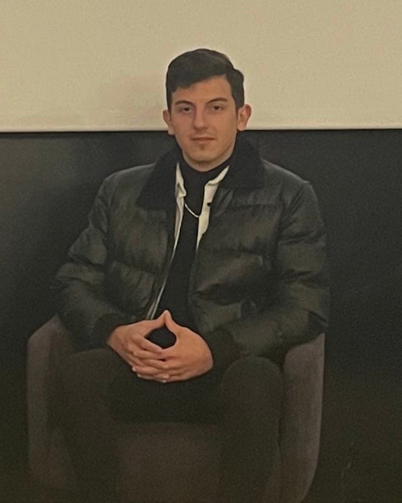 Yunus Emre SAK, kollarını kavuşturmuş, gri beton duvar önünde gülümsüyor